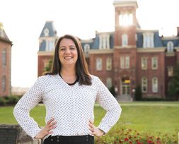 Smiling woman stands with her hands on her hips with Woodburn Hall and the sun shining in the background. She wears a polka dot white blouse, black dress pants and has dark hair. 